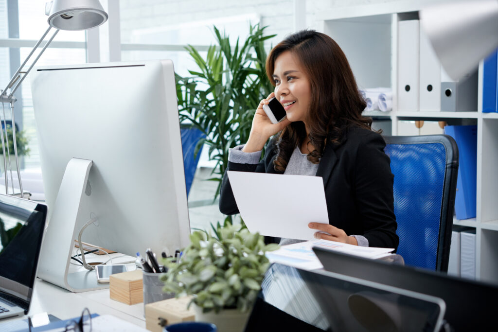 Woman in a black blazer talking on a smartphone at a desk with papers, a computer monitor, and a plant in an bright office setting